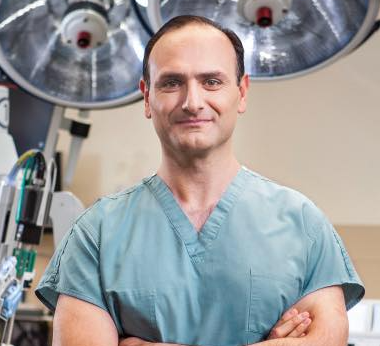 Dr Samir Hage wearing blue scrubs and standing in front of the robotic surgery machine